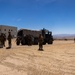 A group of military personnel in combat gear are gathered around a large canvas-covered military truck in an arid desert training environment. The scene appears to be a tactical exercise or training operation at a military installation, featuring mock urban structures in the background. The setting is consistent with a desert training facility, likely in the American Southwest based on terrain and architecture.