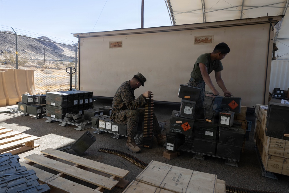 Two military personnel, appearing to be U.S. Marines based on camouflage uniforms, are working with ammunition and military supply crates in an outdoor covered area in a desert environment. One Marine is seated handling a belt of linked ammunition, while the other is standing and organizing metal ammunition cans stacked on pallets. The setting appears to be a forward operating base or training facility, with chain-link fencing, barbed wire, and desert mountains visible in the background.