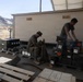 Two military personnel, appearing to be U.S. Marines based on camouflage uniforms, are working with ammunition and military supply crates in an outdoor covered area in a desert environment. One Marine is seated handling a belt of linked ammunition, while the other is standing and organizing metal ammunition cans stacked on pallets. The setting appears to be a forward operating base or training facility, with chain-link fencing, barbed wire, and desert mountains visible in the background.