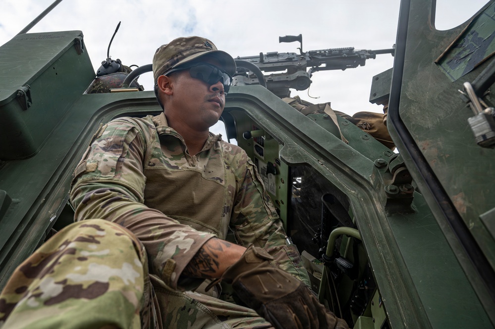 A military soldier in MultiCam/OCP camouflage uniform is positioned in or on a military vehicle, looking outward with a focused expression. A crew-served machine gun (appears to be an M240 or similar) is mounted above and behind him on the vehicle. The low-angle photograph emphasizes the soldier's alert posture and the tactical equipment surrounding him.