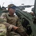 A military soldier in MultiCam/OCP camouflage uniform is positioned in or on a military vehicle, looking outward with a focused expression. A crew-served machine gun (appears to be an M240 or similar) is mounted above and behind him on the vehicle. The low-angle photograph emphasizes the soldier's alert posture and the tactical equipment surrounding him.