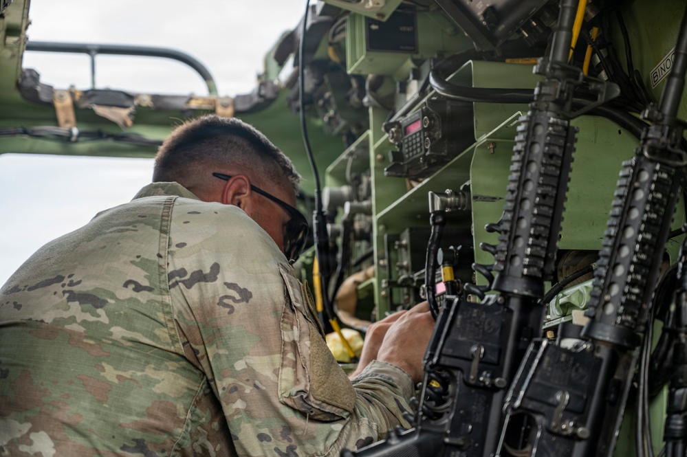 A military service member in OCP (Operational Camouflage Pattern) uniform is working inside or alongside a military vehicle, interacting with equipment and controls. The individual appears to be performing maintenance, configuration, or operational tasks on vehicle-mounted systems. Multiple weapons and communication devices are visible in the immediate workspace.