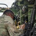 A military service member in OCP (Operational Camouflage Pattern) uniform is working inside or alongside a military vehicle, interacting with equipment and controls. The individual appears to be performing maintenance, configuration, or operational tasks on vehicle-mounted systems. Multiple weapons and communication devices are visible in the immediate workspace.