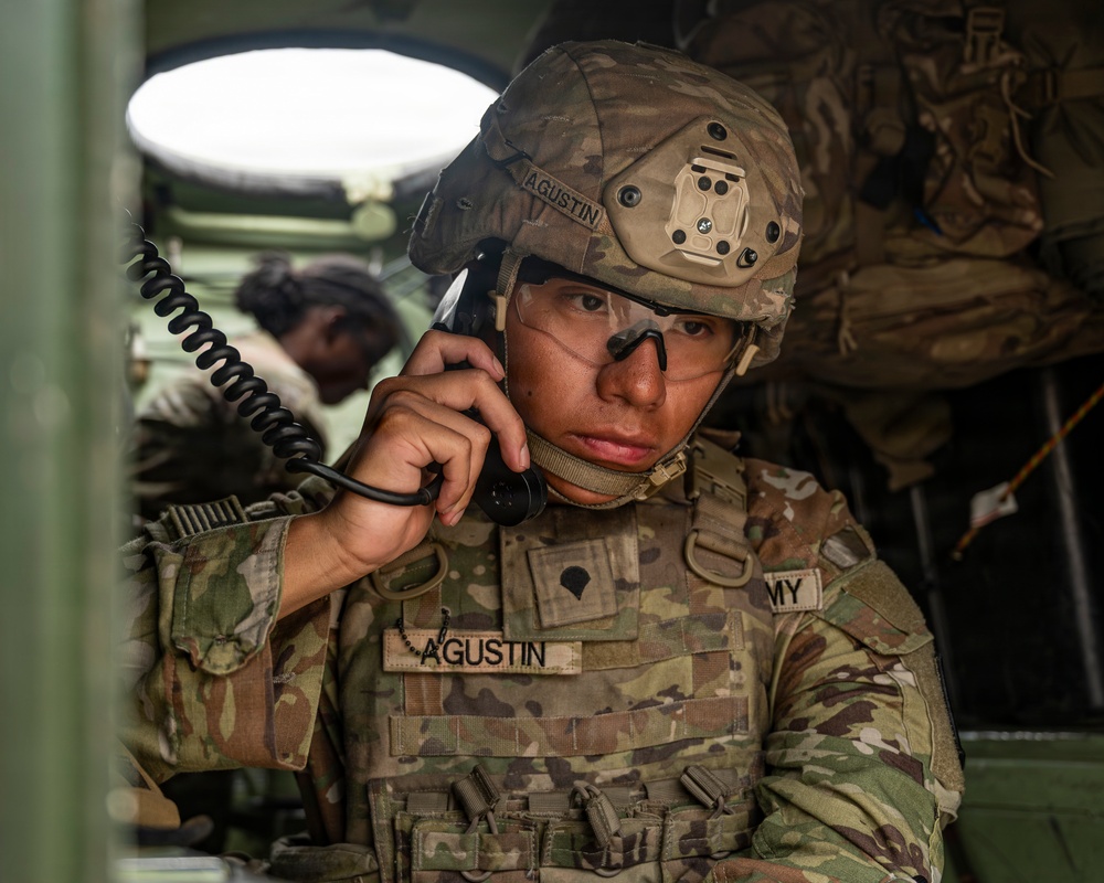 A U.S. Army soldier identified as 'Agustin' is using a military radio handset inside what appears to be a tactical vehicle or command post. The soldier is wearing full combat gear including an OCP (Operational Camouflage Pattern) uniform, tactical helmet with a night vision mount, and body armor. A second soldier is visible in the background.