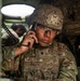 A U.S. Army soldier identified as 'Agustin' is using a military radio handset inside what appears to be a tactical vehicle or command post. The soldier is wearing full combat gear including an OCP (Operational Camouflage Pattern) uniform, tactical helmet with a night vision mount, and body armor. A second soldier is visible in the background.