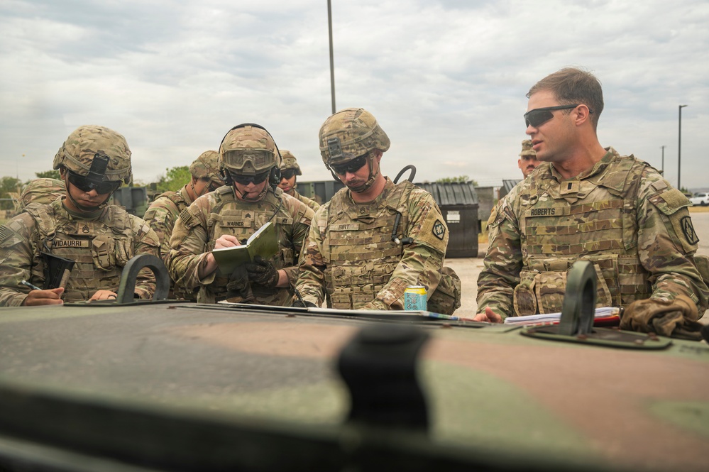 A group of U.S. Army soldiers in OCP (Operational Camouflage Pattern) uniforms are gathered around the hood of a military vehicle, reviewing documents and maps. At least four soldiers are identifiable by name tapes: Vidaurri, Warner, Birt, and Roberts. The scene appears to be a planning or briefing session conducted outdoors at a military installation.