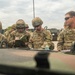 A group of U.S. Army soldiers in OCP (Operational Camouflage Pattern) uniforms are gathered around the hood of a military vehicle, reviewing documents and maps. At least four soldiers are identifiable by name tapes: Vidaurri, Warner, Birt, and Roberts. The scene appears to be a planning or briefing session conducted outdoors at a military installation.