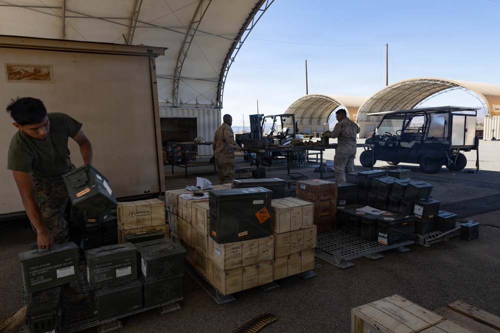 Military personnel in camouflage uniforms are handling and organizing ammunition crates and supply boxes at what appears to be a military logistics or ordnance staging area. The scene shows multiple pallets loaded with wooden and metal ammunition cans under large arched tent structures. The setting appears to be a desert or arid environment based on the terrain and sky visible in the background.