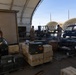 Military personnel in camouflage uniforms are handling and organizing ammunition crates and supply boxes at what appears to be a military logistics or ordnance staging area. The scene shows multiple pallets loaded with wooden and metal ammunition cans under large arched tent structures. The setting appears to be a desert or arid environment based on the terrain and sky visible in the background.