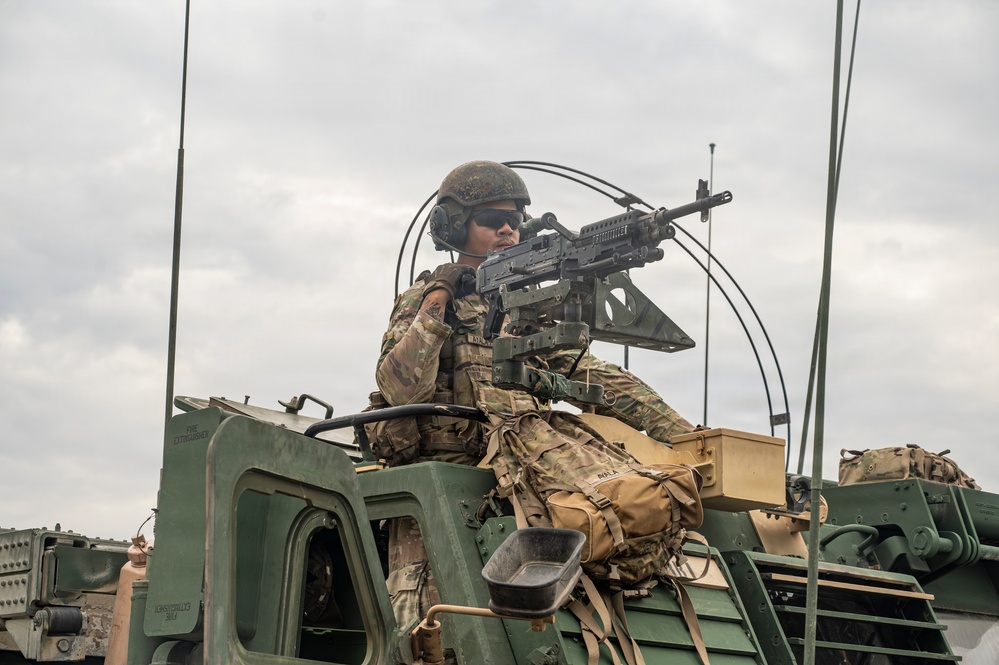 A soldier in full combat gear is positioned in a turret hatch atop a military armored vehicle, manning what appears to be an M249 or M240 machine gun. The soldier is wearing OCP (Operational Camouflage Pattern) uniform, a combat helmet, communications headset, and tactical sunglasses. The image appears to be taken during a training exercise or operational deployment.