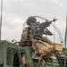 A soldier in full combat gear is positioned in a turret hatch atop a military armored vehicle, manning what appears to be an M249 or M240 machine gun. The soldier is wearing OCP (Operational Camouflage Pattern) uniform, a combat helmet, communications headset, and tactical sunglasses. The image appears to be taken during a training exercise or operational deployment.