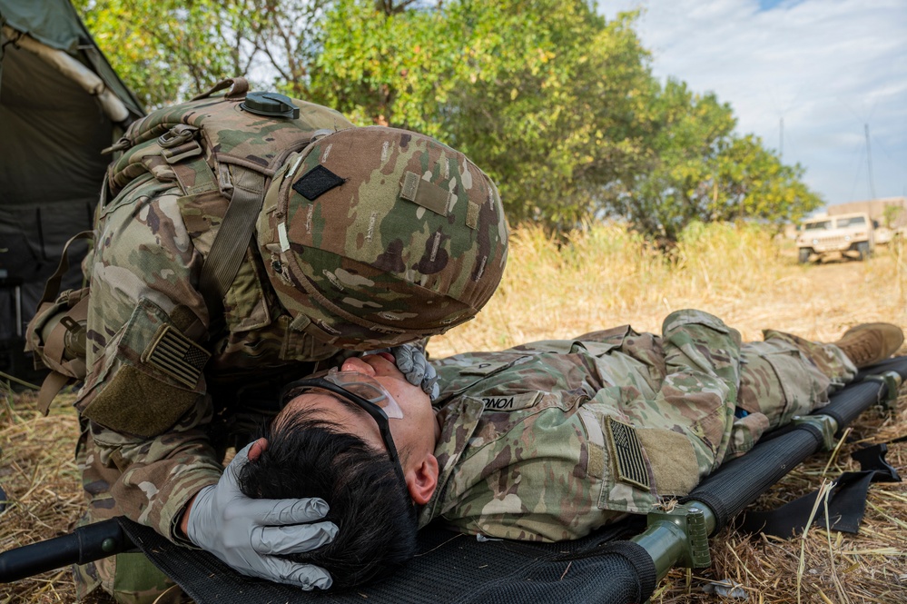 A U.S. Army soldier in full combat gear and gloves is performing a medical assessment or airway check on a fellow soldier lying on a litter/stretcher on the ground. The scene appears to be a field medical training exercise, with a military tent visible on the left and a military vehicle in the background. The prone soldier has medical tape applied to their face, consistent with a simulated casualty scenario.