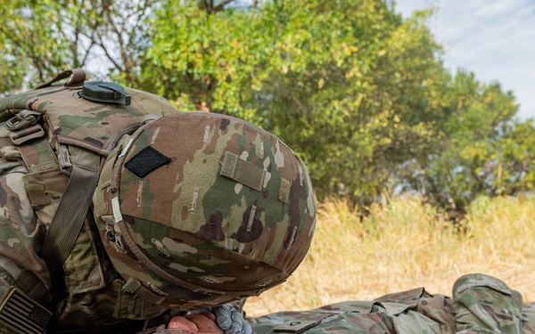 A U.S. Army soldier in full combat gear and gloves is performing a medical assessment or airway check on a fellow soldier lying on a litter/stretcher on the ground. The scene appears to be a field medical training exercise, with a military tent visible on the left and a military vehicle in the background. The prone soldier has medical tape applied to their face, consistent with a simulated casualty scenario.