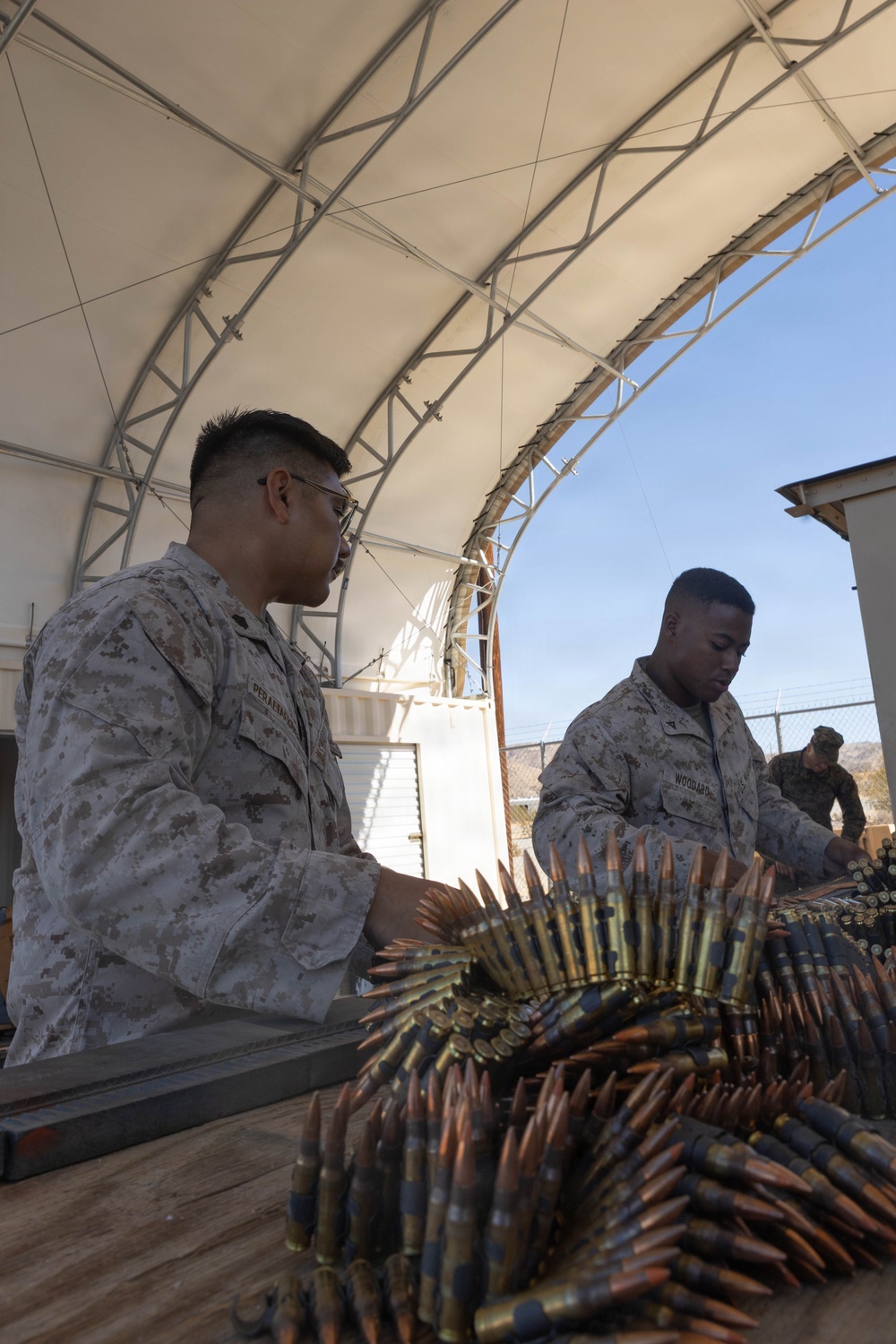 Two U.S. Marines in MARPAT desert camouflage uniforms are handling large quantities of linked ammunition at a table under an open-sided tent structure. A third Marine is visible in the background near a chain-link fence. The setting appears to be a military installation in an arid environment, likely an ammunition point or range support area.