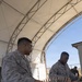 Two U.S. Marines in MARPAT desert camouflage uniforms are handling large quantities of linked ammunition at a table under an open-sided tent structure. A third Marine is visible in the background near a chain-link fence. The setting appears to be a military installation in an arid environment, likely an ammunition point or range support area.