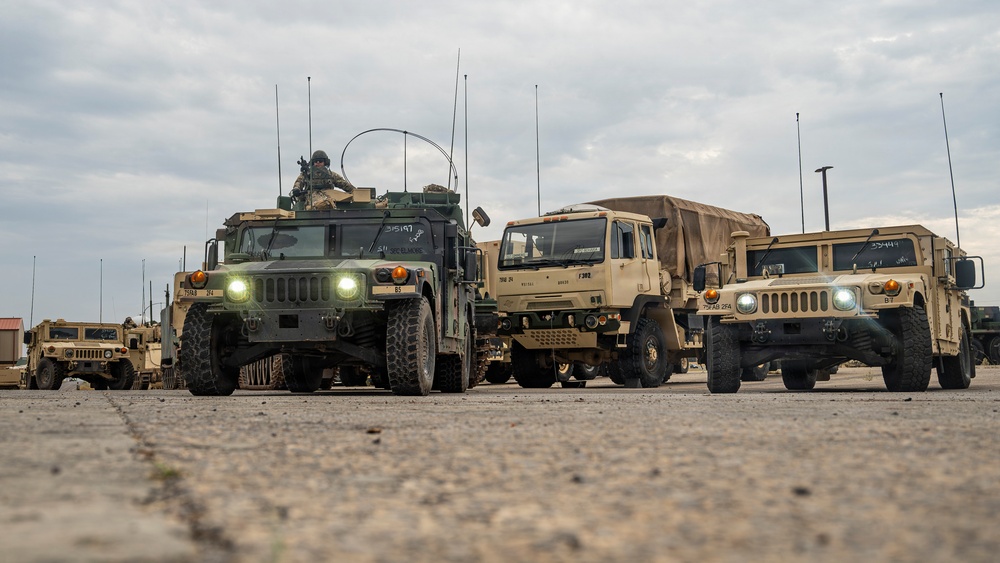 A convoy of U.S. military tactical vehicles is staged or moving on a paved surface under overcast skies. The formation includes multiple HMMWVs (Humvees) and at least one LMTV (Light Medium Tactical Vehicle) with a canvas-covered cargo bed. At least one soldier is visible manning a turret position on the lead Humvee.