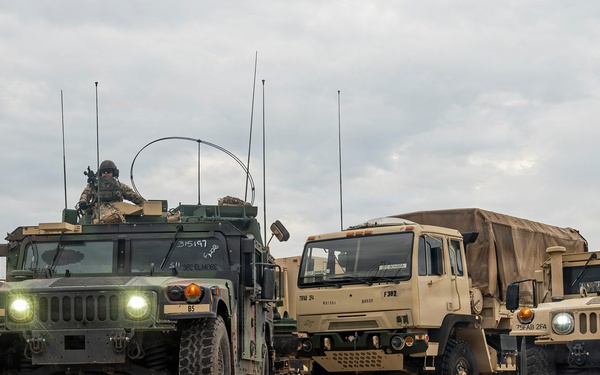 A convoy of U.S. military tactical vehicles is staged or moving on a paved surface under overcast skies. The formation includes multiple HMMWVs (Humvees) and at least one LMTV (Light Medium Tactical Vehicle) with a canvas-covered cargo bed. At least one soldier is visible manning a turret position on the lead Humvee.