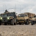 A convoy of U.S. military tactical vehicles is staged or moving on a paved surface under overcast skies. The formation includes multiple HMMWVs (Humvees) and at least one LMTV (Light Medium Tactical Vehicle) with a canvas-covered cargo bed. At least one soldier is visible manning a turret position on the lead Humvee.