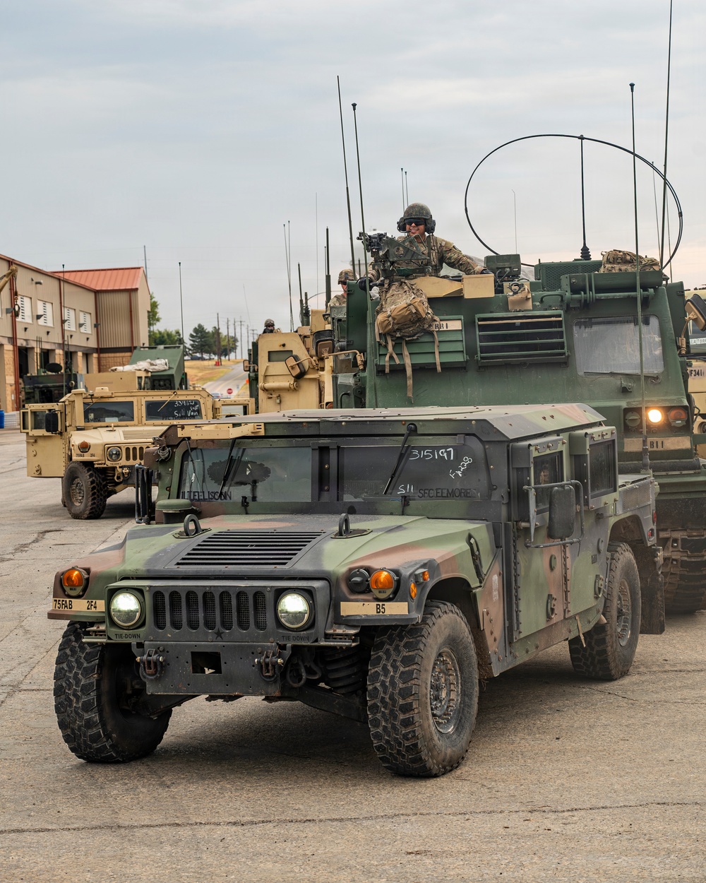 A military convoy of HMMWV (Humvee) vehicles and at least one larger tactical vehicle is staged or moving through what appears to be a military installation or base. Soldiers in camouflage uniforms and helmets are visible, with one manning a weapon system on top of a vehicle. Vehicle markings including '75FAB', '2F4', 'B5', '315197', and 'SFC ELMORE' are visible on the lead vehicles.