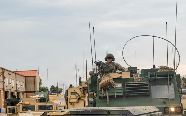 A military convoy of HMMWV (Humvee) vehicles and at least one larger tactical vehicle is staged or moving through what appears to be a military installation or base. Soldiers in camouflage uniforms and helmets are visible, with one manning a weapon system on top of a vehicle. Vehicle markings including '75FAB', '2F4', 'B5', '315197', and 'SFC ELMORE' are visible on the lead vehicles.