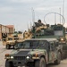 A military convoy of HMMWV (Humvee) vehicles and at least one larger tactical vehicle is staged or moving through what appears to be a military installation or base. Soldiers in camouflage uniforms and helmets are visible, with one manning a weapon system on top of a vehicle. Vehicle markings including '75FAB', '2F4', 'B5', '315197', and 'SFC ELMORE' are visible on the lead vehicles.