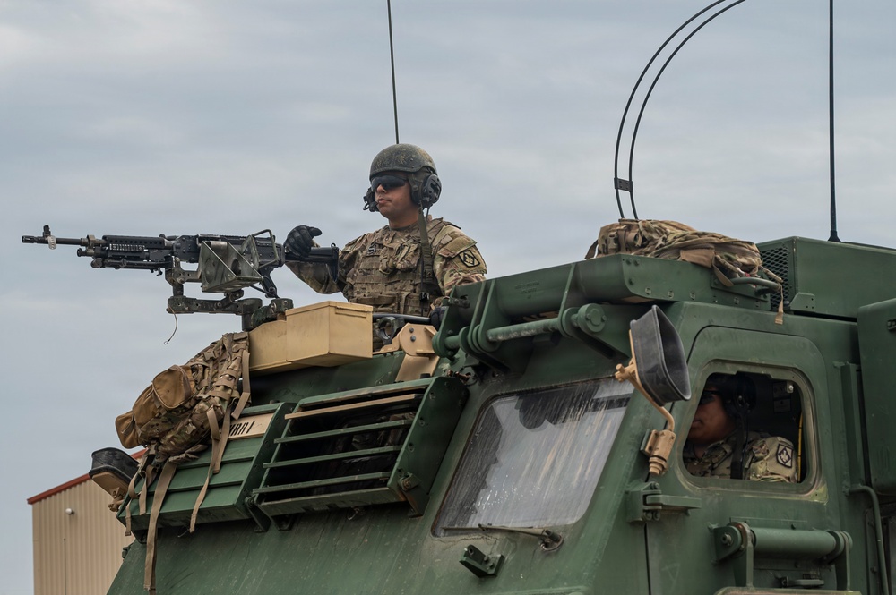 Two military personnel are positioned on top of and inside a large green armored military vehicle. One soldier is manning a mounted machine gun from a turret position on top of the vehicle, while another is visible through the cab window. The soldiers are wearing full combat gear including helmets, tactical headsets, and camouflage uniforms.