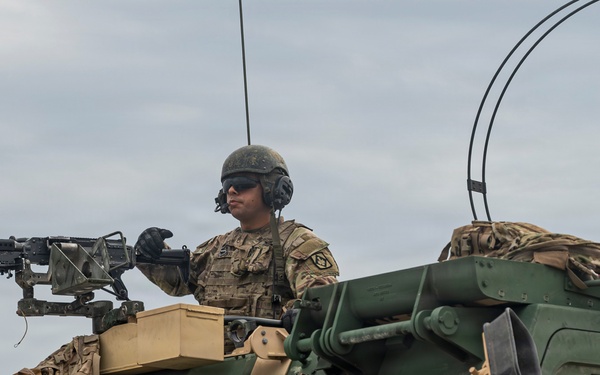 Two military personnel are positioned on top of and inside a large green armored military vehicle. One soldier is manning a mounted machine gun from a turret position on top of the vehicle, while another is visible through the cab window. The soldiers are wearing full combat gear including helmets, tactical headsets, and camouflage uniforms.