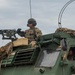 Two military personnel are positioned on top of and inside a large green armored military vehicle. One soldier is manning a mounted machine gun from a turret position on top of the vehicle, while another is visible through the cab window. The soldiers are wearing full combat gear including helmets, tactical headsets, and camouflage uniforms.