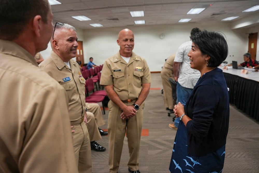 A group of U.S. Navy personnel in tan service uniforms are engaged in conversation with a civilian woman in a navy blue cardigan and patterned skirt. The setting appears to be a conference or meeting room with red chairs and a long table visible in the background. The interaction appears cordial and professional in nature.