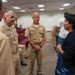 A group of U.S. Navy personnel in tan service uniforms are engaged in conversation with a civilian woman in a navy blue cardigan and patterned skirt. The setting appears to be a conference or meeting room with red chairs and a long table visible in the background. The interaction appears cordial and professional in nature.