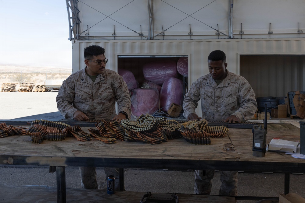 Two U.S. Marines in desert MARPAT camouflage uniforms are working at an outdoor table handling linked ammunition belts. They appear to be sorting, counting, or preparing machine gun ammunition at what looks like a forward operating base or training facility. A shipping container and desert terrain are visible in the background.