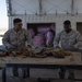 Two U.S. Marines in desert MARPAT camouflage uniforms are working at an outdoor table handling linked ammunition belts. They appear to be sorting, counting, or preparing machine gun ammunition at what looks like a forward operating base or training facility. A shipping container and desert terrain are visible in the background.