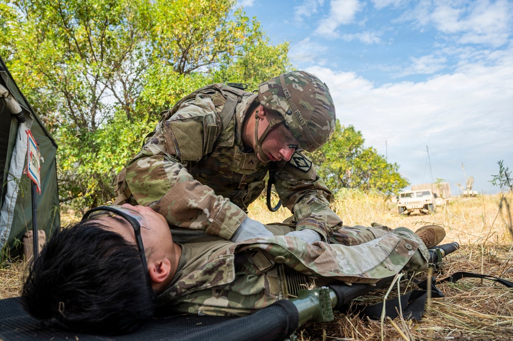 A U.S. Army soldier in full OCP (Operational Camouflage Pattern) uniform and combat gear is performing medical treatment on a simulated casualty lying on a litter/stretcher. The scene appears to be a field medical training exercise, with the patient wearing a non-rebreather oxygen mask. Military vehicles and a tent are visible in the background.