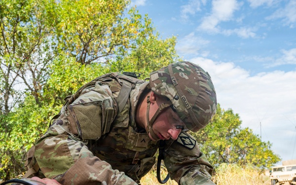A U.S. Army soldier in full OCP (Operational Camouflage Pattern) uniform and combat gear is performing medical treatment on a simulated casualty lying on a litter/stretcher. The scene appears to be a field medical training exercise, with the patient wearing a non-rebreather oxygen mask. Military vehicles and a tent are visible in the background.