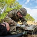 A U.S. Army soldier in full OCP (Operational Camouflage Pattern) uniform and combat gear is performing medical treatment on a simulated casualty lying on a litter/stretcher. The scene appears to be a field medical training exercise, with the patient wearing a non-rebreather oxygen mask. Military vehicles and a tent are visible in the background.
