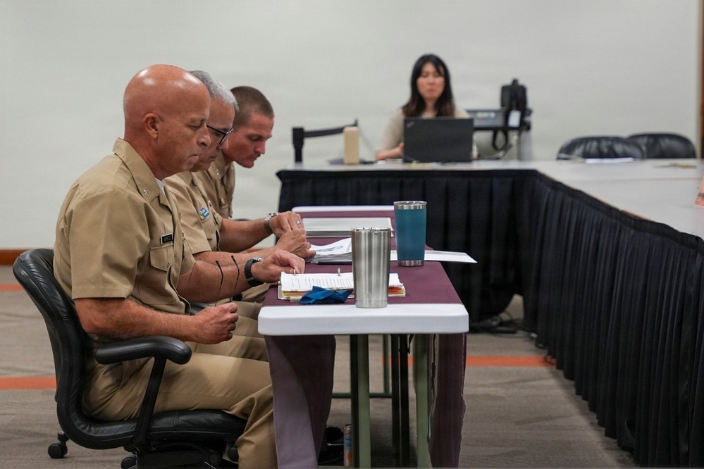 Three individuals in U.S. Navy khaki uniforms are seated at a table reviewing documents during what appears to be a formal military meeting or board proceeding. In the background, a civilian woman is working at a laptop, likely serving as a recorder or administrative support. The setting appears to be a conference or meeting room with draped tables arranged in a formal layout.
