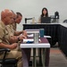 Three individuals in U.S. Navy khaki uniforms are seated at a table reviewing documents during what appears to be a formal military meeting or board proceeding. In the background, a civilian woman is working at a laptop, likely serving as a recorder or administrative support. The setting appears to be a conference or meeting room with draped tables arranged in a formal layout.