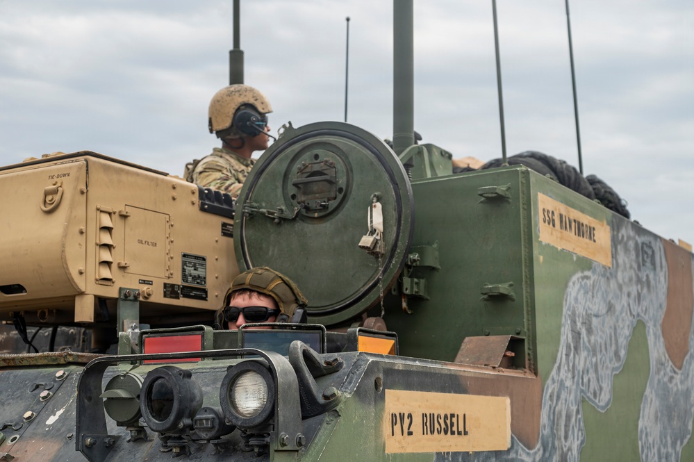 Two soldiers are positioned in or on a military armored vehicle, likely a Bradley Fighting Vehicle or similar tracked platform. The vehicle displays name placards reading 'PY2 RUSSELL' and 'SSG HAWTHORNE', indicating crew assignments. The scene appears to be a field training exercise or operational deployment.