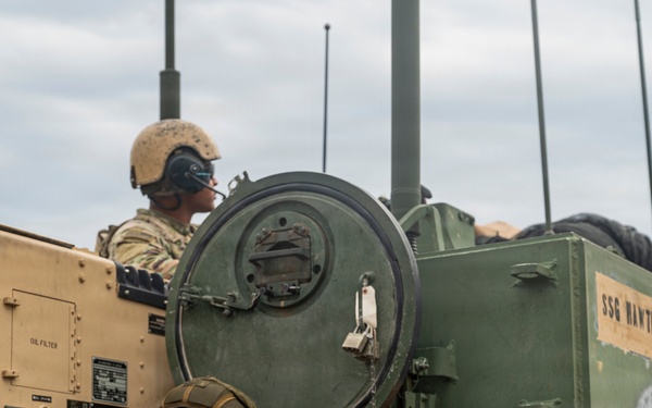 Two soldiers are positioned in or on a military armored vehicle, likely a Bradley Fighting Vehicle or similar tracked platform. The vehicle displays name placards reading 'PY2 RUSSELL' and 'SSG HAWTHORNE', indicating crew assignments. The scene appears to be a field training exercise or operational deployment.