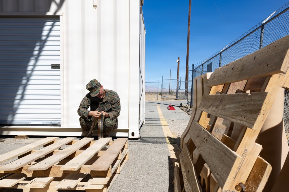 A military service member in camouflage uniform is crouched on wooden pallets between two white storage containers, handling what appears to be an ammunition belt. The setting appears to be a military installation or training facility in an arid desert environment, with chain-link fencing and utility poles visible in the background. The scene suggests ammunition preparation or inspection activities.