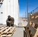 A military service member in camouflage uniform is crouched on wooden pallets between two white storage containers, handling what appears to be an ammunition belt. The setting appears to be a military installation or training facility in an arid desert environment, with chain-link fencing and utility poles visible in the background. The scene suggests ammunition preparation or inspection activities.