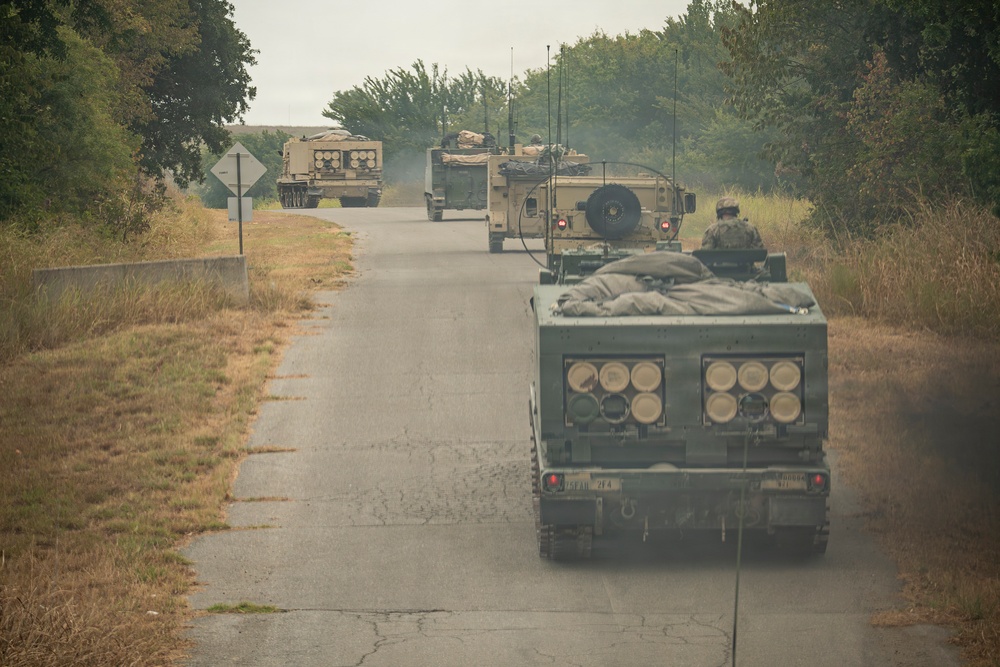 A military convoy of multiple tracked vehicles is traveling along a narrow paved road flanked by dry grass and trees. The vehicles appear to be M270 Multiple Launch Rocket Systems (MLRS) or similar artillery rocket systems, identifiable by their distinctive launcher pod configurations visible on the rear vehicle. At least one soldier is visible manning a position on one of the vehicles.