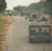 A military convoy of multiple tracked vehicles is traveling along a narrow paved road flanked by dry grass and trees. The vehicles appear to be M270 Multiple Launch Rocket Systems (MLRS) or similar artillery rocket systems, identifiable by their distinctive launcher pod configurations visible on the rear vehicle. At least one soldier is visible manning a position on one of the vehicles.