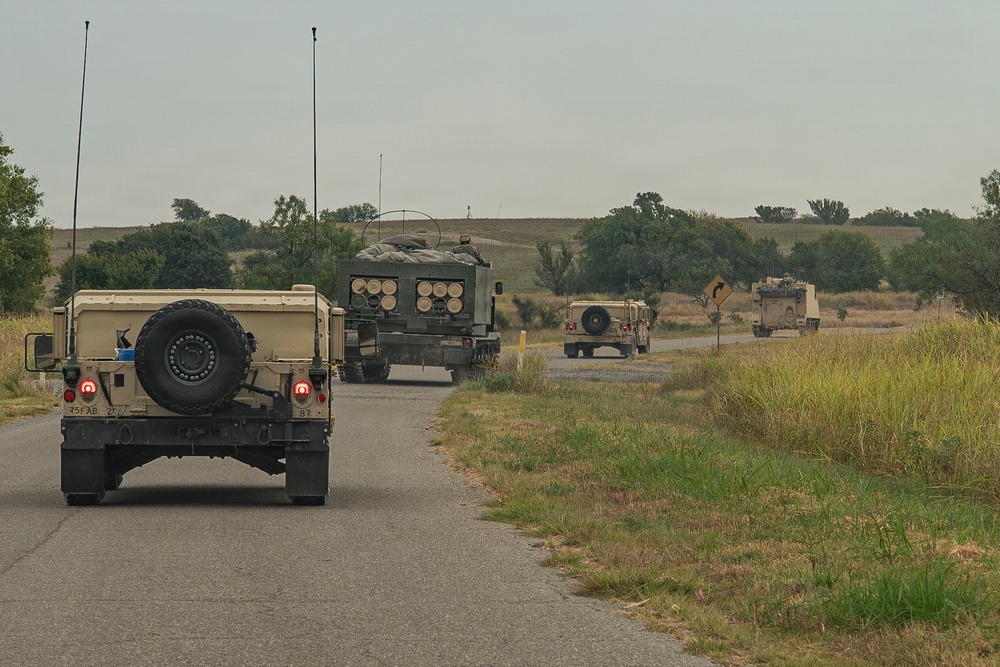 A military convoy of at least four vehicles is traveling along a two-lane rural road through open grassland terrain. The convoy includes HMMWV (Humvee) vehicles and what appears to be an M270 Multiple Launch Rocket System (MLRS) or similar tracked artillery vehicle. The vehicles are in desert tan/olive drab camouflage coloring, consistent with U.S. Army equipment.