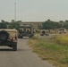A military convoy of at least four vehicles is traveling along a two-lane rural road through open grassland terrain. The convoy includes HMMWV (Humvee) vehicles and what appears to be an M270 Multiple Launch Rocket System (MLRS) or similar tracked artillery vehicle. The vehicles are in desert tan/olive drab camouflage coloring, consistent with U.S. Army equipment.