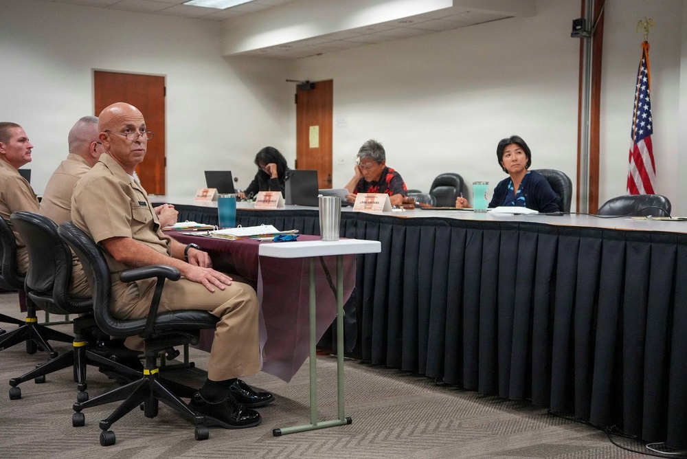 A formal meeting or hearing is taking place in a conference room setting. Military personnel in tan/khaki uniforms are seated on one side, facing a panel of civilian officials seated behind a draped table. An American flag is visible in the background, suggesting an official government or military-civilian engagement.