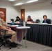 A formal meeting or hearing is taking place in a conference room setting. Military personnel in tan/khaki uniforms are seated on one side, facing a panel of civilian officials seated behind a draped table. An American flag is visible in the background, suggesting an official government or military-civilian engagement.