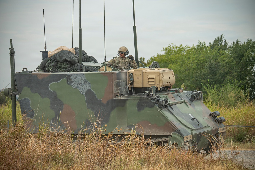 A military armored command vehicle, likely an M577 or similar command post carrier variant, is positioned in a field environment with a soldier visible in the open hatch. The vehicle displays a three-color NATO camouflage pattern (green, brown, black) and is equipped with multiple radio antennas, indicating a command and control role. The soldier appears to be communicating via radio headset while monitoring the surrounding area.