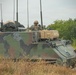 A military armored command vehicle, likely an M577 or similar command post carrier variant, is positioned in a field environment with a soldier visible in the open hatch. The vehicle displays a three-color NATO camouflage pattern (green, brown, black) and is equipped with multiple radio antennas, indicating a command and control role. The soldier appears to be communicating via radio headset while monitoring the surrounding area.