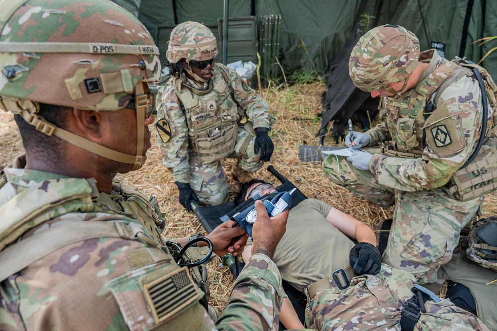 U.S. Army soldiers are conducting a field medical training exercise, treating a simulated casualty on a litter in an outdoor environment near a military tent. Multiple personnel are performing coordinated medical tasks including airway management and documentation. Soldiers are wearing OCP (Operational Camouflage Pattern) uniforms with unit patches visible, including a 1st Cavalry Division patch.
